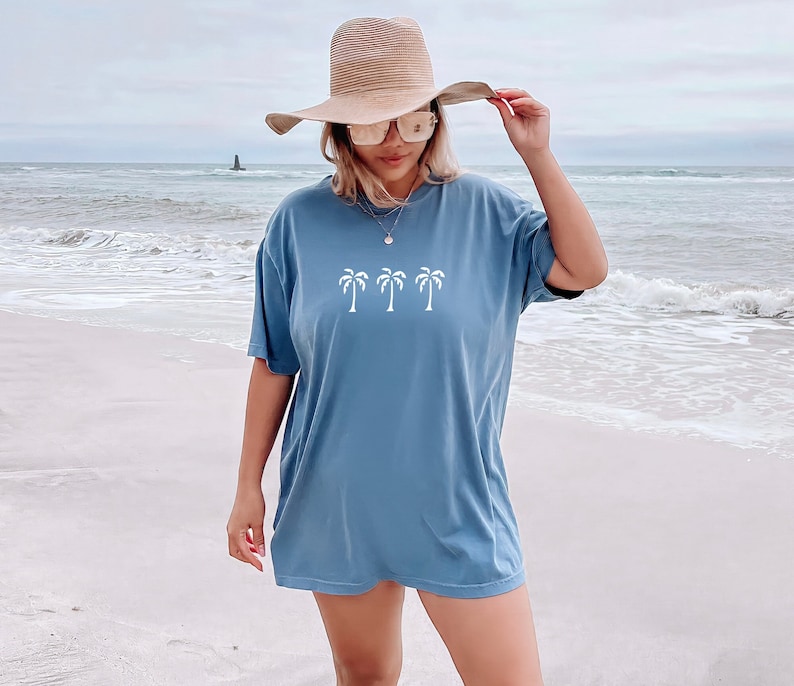 Puede incluir: Una mujer lleva una camiseta azul de gran tama&ntilde;o con un gr&aacute;fico de palmeras blancas en la parte delantera. Lleva un sombrero de paja de ala ancha y gafas de sol. La mujer est&aacute; de pie en una playa de arena.