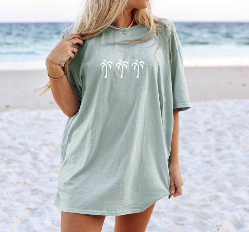 Puede incluir: Una camiseta de gran tama&ntilde;o de color azul claro con un gr&aacute;fico de cuatro palmeras blancas. La camiseta la lleva puesta una persona en una playa.