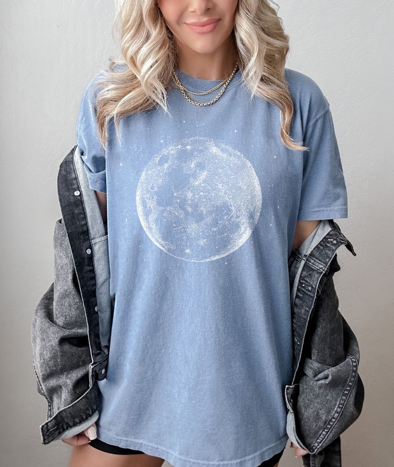 May include: A pale blue t-shirt with a white illustration of the moon. The celestial design is centered on the shirt, with a starry background. The shirt is paired with a dark denim jacket.