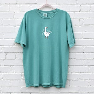 May include: Teal blue short-sleeved t-shirt with a white goose graphic and the text "Look at me I'm quacking".