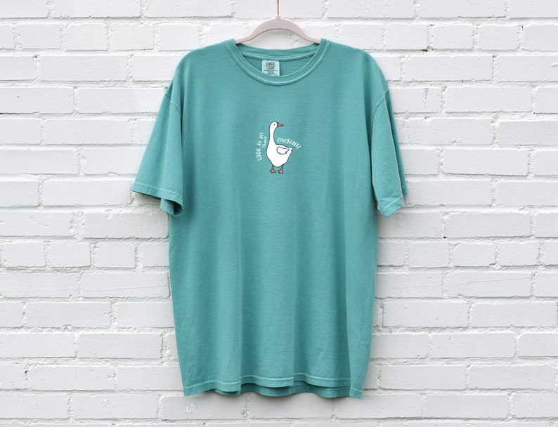 May include: Teal blue short-sleeved t-shirt with a white goose graphic and the text "Look at me I'm quacking".