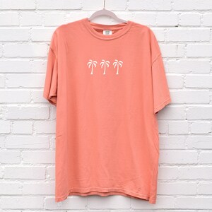 Puede incluir: Una camiseta de color coral con un gr&aacute;fico blanco de cuatro palmeras. La camiseta tiene cuello redondo y mangas cortas.