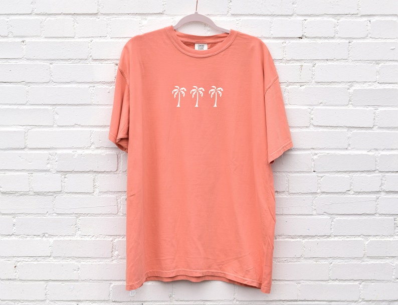 Puede incluir: Una camiseta de color coral con un gr&aacute;fico blanco de cuatro palmeras. La camiseta tiene cuello redondo y mangas cortas.