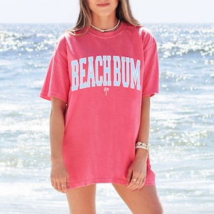 以下が含まれることがあります： ビーチで立っている人の着ている、ピンク色のオーバーサイズのTシャツ。Tシャツには薄い青色の「BEACH BUM」の文字と、その下にヤシの木のグラフィックが描かれている。