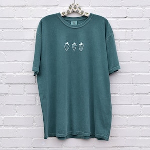 Op de afbeelding: Een tealblauw t-shirt met een wit geborduurd ontwerp van drie aardbeien.