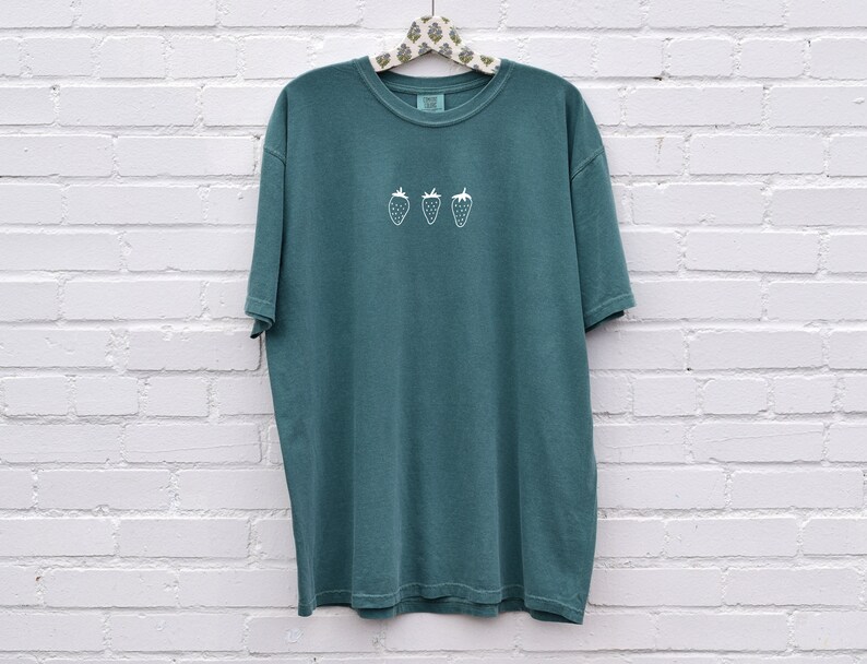 Op de afbeelding: Een tealblauw t-shirt met een wit geborduurd ontwerp van drie aardbeien.