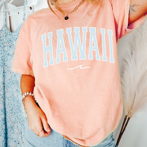 May include: A peach-colored t-shirt with the word "HAWAII" printed in light blue on the front. The shirt has a relaxed fit and a crew neck.