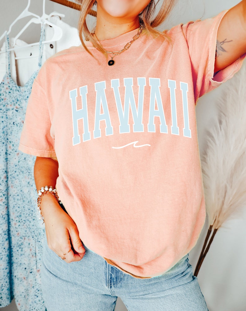May include: A peach-colored t-shirt with the word "HAWAII" printed in light blue on the front. The shirt has a relaxed fit and a crew neck.