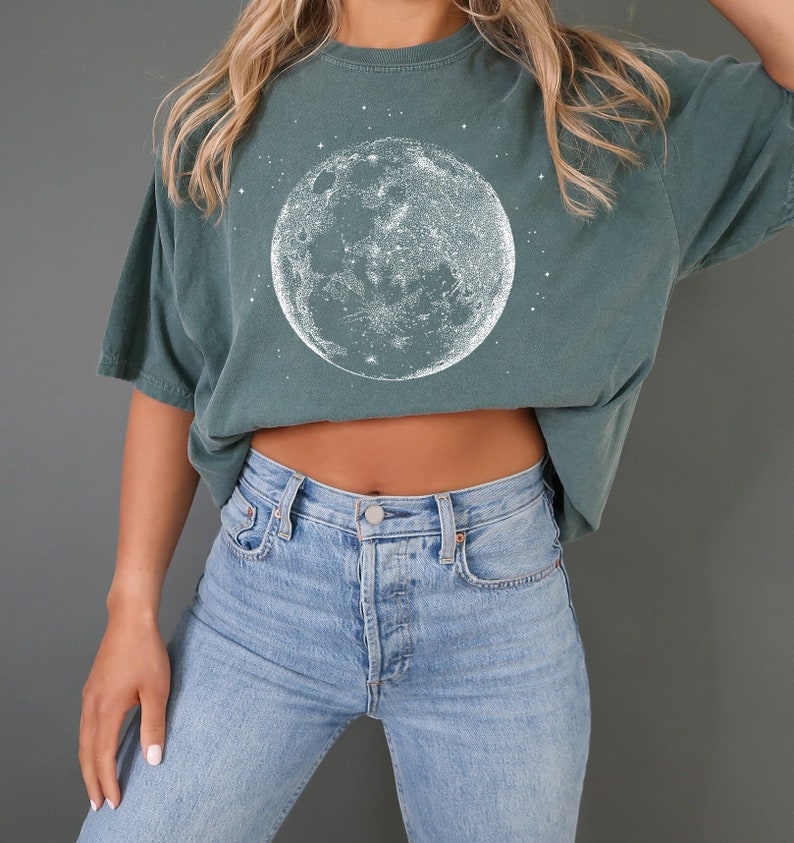 May include: A teal t-shirt with a white illustration of the moon and stars. The moon graphic is detailed. The shirt is worn with light blue denim jeans.