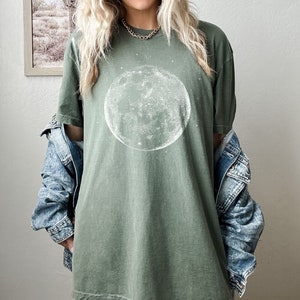 May include: An olive green t-shirt with a white moon and star design. The t-shirt has a loose fit and is made of soft fabric. A denim jacket is worn over the t-shirt. This is a casual, everyday wear item.