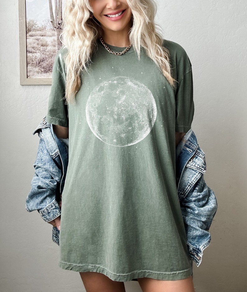 May include: An olive green t-shirt with a white moon and star design. The t-shirt has a loose fit and is made of soft fabric. A denim jacket is worn over the t-shirt. This is a casual, everyday wear item.