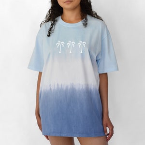 Puede incluir: Una camiseta azul claro tie-dye con un gr&aacute;fico de palmera blanca. La camiseta tiene un dise&ntilde;o degradado, que se desvanece de azul claro a azul m&aacute;s oscuro.