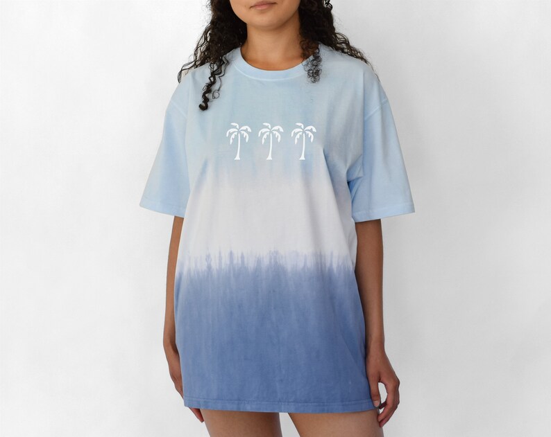 Puede incluir: Una camiseta azul claro tie-dye con un gr&aacute;fico de palmera blanca. La camiseta tiene un dise&ntilde;o degradado, que se desvanece de azul claro a azul m&aacute;s oscuro.