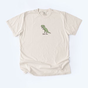 May include: A beige t-shirt with a green cartoon dinosaur and the text "bite me" printed on the front.