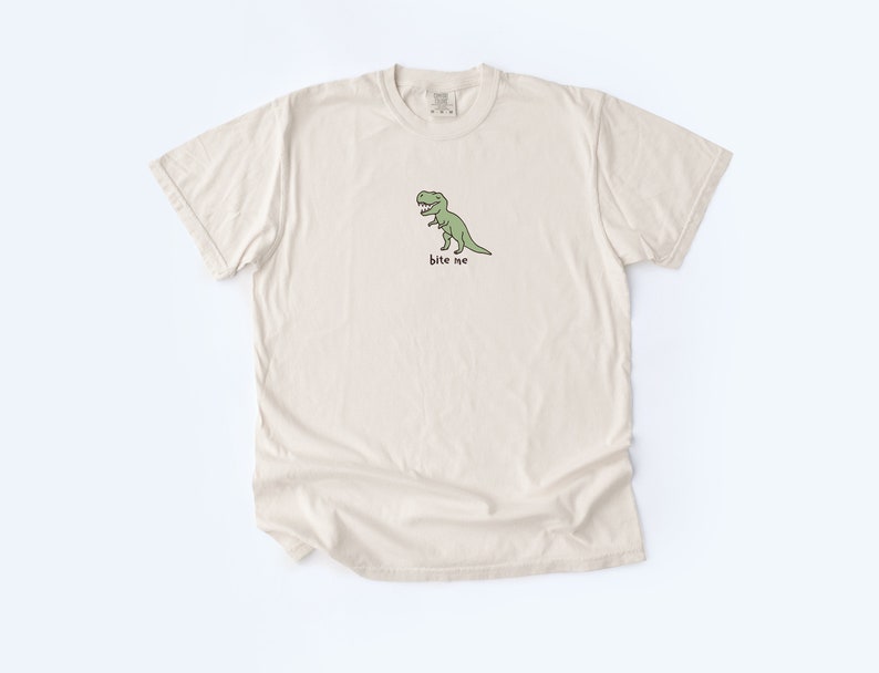 May include: A beige t-shirt with a green cartoon dinosaur and the text "bite me" printed on the front.