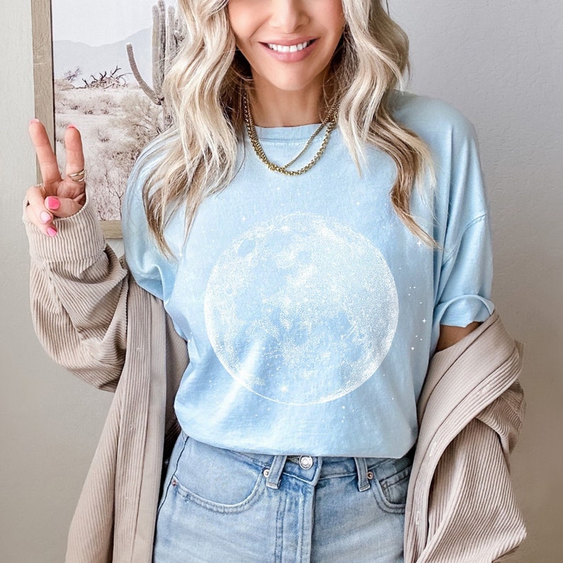 May include: Pale blue t-shirt with a large white moon design. The shirt is worn with blue jeans. A beige jacket is draped over the shoulders. A gold necklace is visible. The image has a casual, relaxed feel.