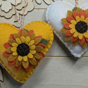 May include: Two heart-shaped ornaments, one yellow and one white, each adorned with a felt sunflower. The sunflowers have layered petals in shades of yellow, orange, and brown, with a dark center. The ornaments hang from twine.