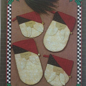 May include: A craft pattern featuring four Santa Claus oven mitts and potholders. The mitts and potholders have red hats, black trim, and cream-colored faces with white beards. The text "Here Comes Santa" is displayed above the images.