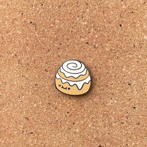 Può includere: Uno spillo smaltato a forma di cornetto alla cannella marrone e bianco con un viso carino. Lo spillo è su uno sfondo di sughero.