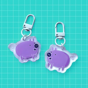 Könnte beinhalten: Zwei Schlüsselanhänger mit einem Cartoon-Schwein-Design. Die Schweine sind lila mit schwarzen Augen und Schnauzen und sind an silberfarbenen Verschlüssen befestigt. Der Hintergrund ist ein türkisfarbenes Gittermuster.