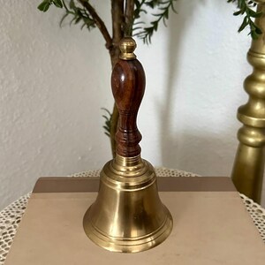May include: A hand bell featuring a polished brass bell base and a dark brown wooden handle with a rounded top. The handle has a turned, grooved design that connects to the metal bell, which has a smooth, flared rim and a shiny, reflective surface.