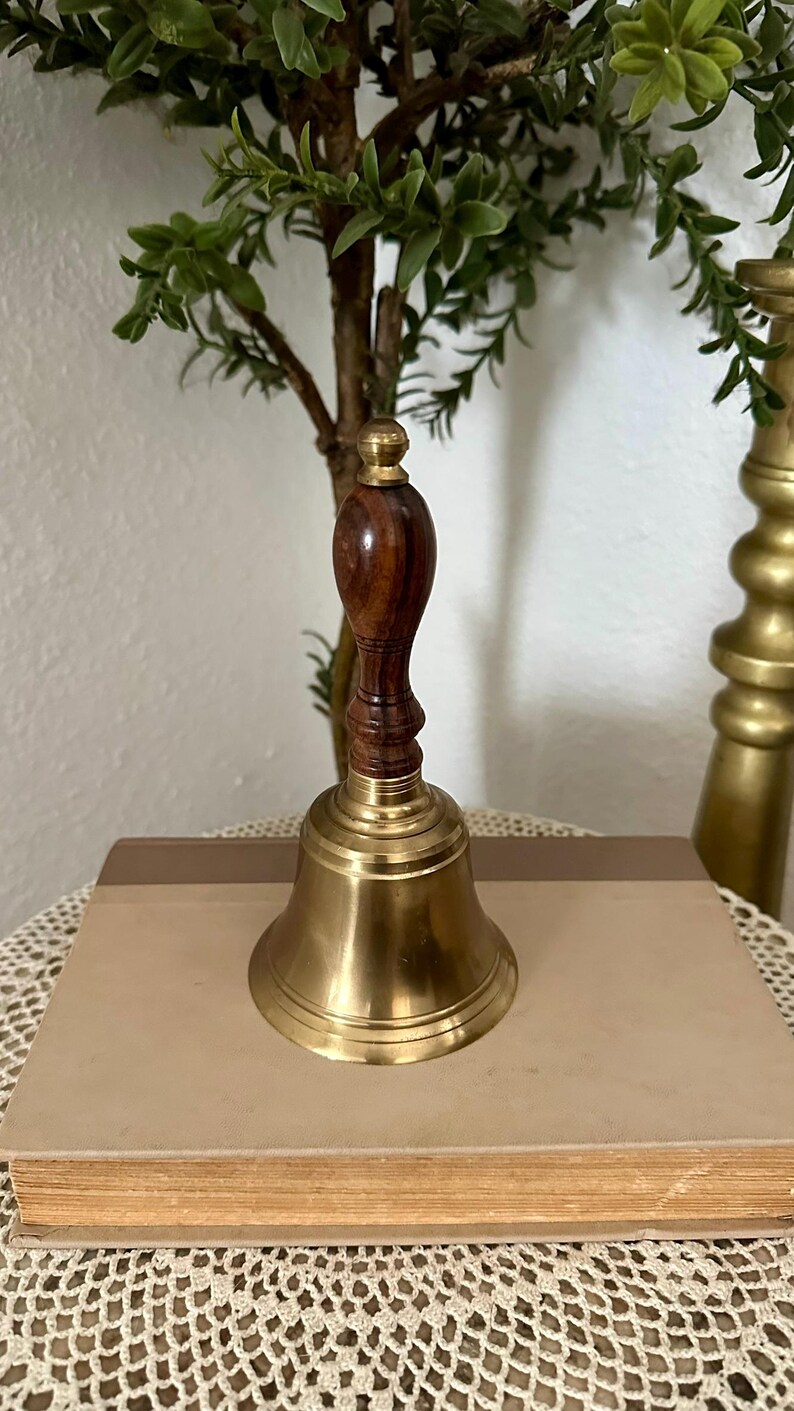 May include: A hand bell featuring a polished brass bell base and a dark brown wooden handle with a rounded top. The handle has a turned, grooved design that connects to the metal bell, which has a smooth, flared rim and a shiny, reflective surface.