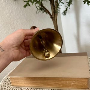 May include: A polished brass hand bell featuring a smooth, flared conical body and a rounded clapper suspended from a twisted metal chain. The bell has a warm, metallic gold finish and a dark wooden handle attached to the top for easy gripping.