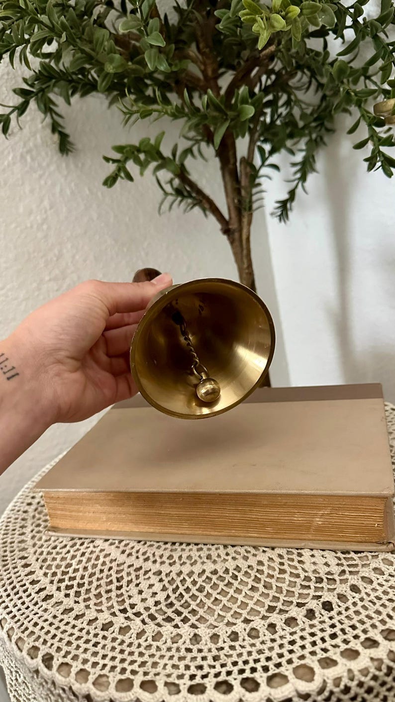 May include: A polished brass hand bell featuring a smooth, flared conical body and a rounded clapper suspended from a twisted metal chain. The bell has a warm, metallic gold finish and a dark wooden handle attached to the top for easy gripping.