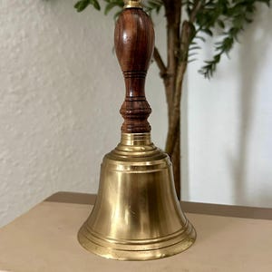 May include: A hand bell featuring a polished brass bell body and a dark brown wooden handle with a rounded top. The handle has a series of horizontal grooves and a smooth, tapered shape. The brass bell has a wide, flared base and a smooth finish.