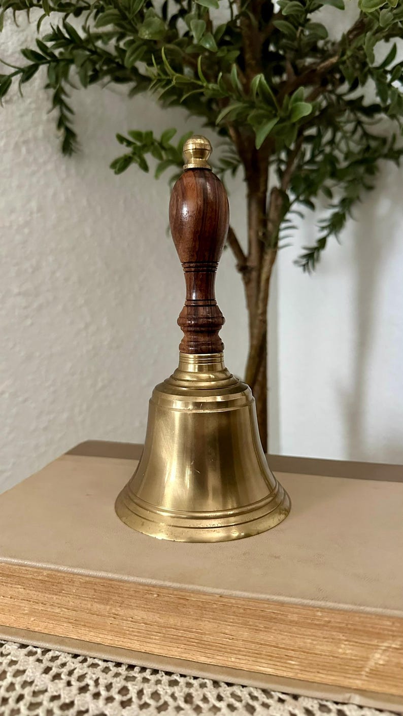 May include: A hand bell featuring a polished brass bell body and a dark brown wooden handle with a rounded top. The handle has a series of horizontal grooves and a smooth, tapered shape. The brass bell has a wide, flared base and a smooth finish.
