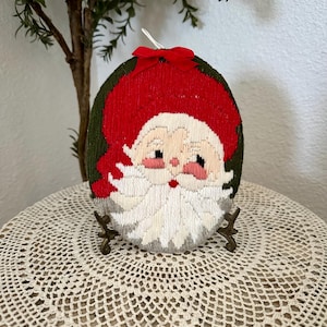 May include: Oval Christmas ornament featuring Santa Claus. The ornament has a red hat and a white beard, with a green border. A small red bow is at the top. The ornament is displayed on a decorative stand.
