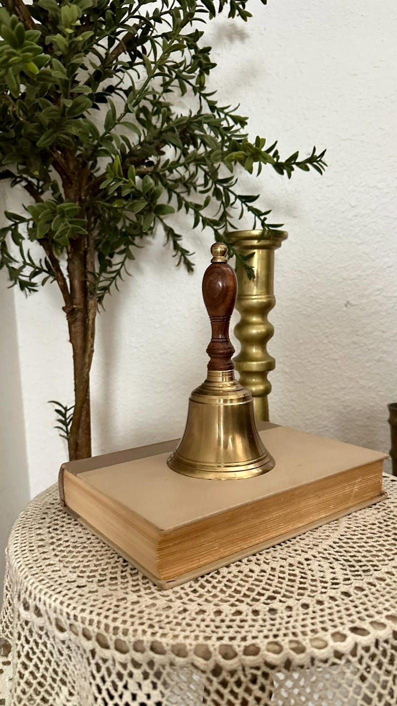 May include: A brass hand bell with a dark brown wooden handle featuring a rounded top knob. The bell has a polished metallic finish and is positioned on top of a rectangular book with a light tan cover, showcasing a classic design and construction.