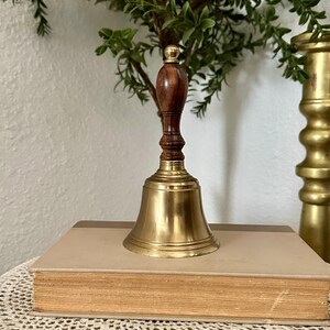 May include: A hand bell featuring a polished brass bell base and a dark brown wooden handle with a rounded top. The bell is positioned upright on top of a closed, rectangular book with a light tan cover and a dark brown spine detail.