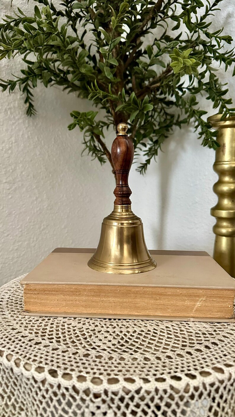 May include: A hand bell featuring a polished brass bell base and a dark brown wooden handle with a rounded top. The bell is positioned upright on top of a closed, rectangular book with a light tan cover and a dark brown spine detail.
