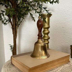 May include: A hand bell featuring a polished brass bell body and a dark brown wooden handle with a rounded top. The bell is positioned on top of a rectangular book with a light tan cover, next to a tall, cylindrical brass candlestick holder.