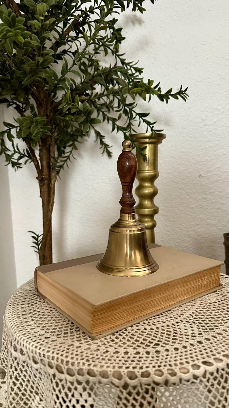 May include: A hand bell featuring a polished brass bell body and a dark brown wooden handle with a rounded top. The bell is positioned on top of a rectangular book with a light tan cover, next to a tall, cylindrical brass candlestick holder.