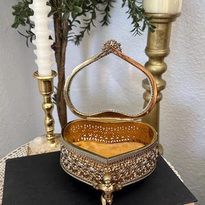 May include: A heart-shaped gold-tone metal trinket box with an open hinged lid, featuring intricate filigree side panels and a small cherub figure at the base. The interior is lined with a soft, mustard-yellow velvet fabric.