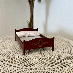 May include: A miniature burgundy bed with a decorative floral design on the headboard. The bed has a white pillow with a bow and is set on a cream-colored crocheted doily. A small tree is in the background.