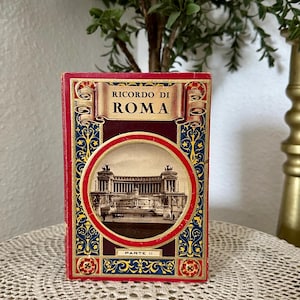 May include: A rectangular book titled Ricordo di Roma Parte II with a red cover featuring a central circular sepia-toned photograph of a large neoclassical building with columns and statues, surrounded by ornate blue and gold decorative patterns.