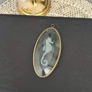 May include: An oval pendant featuring a light blue seahorse silhouette centered on a dark gray, faceted glass background. The pendant is encased in a textured gold-tone metal frame with a small circular bail at the top for attachment.