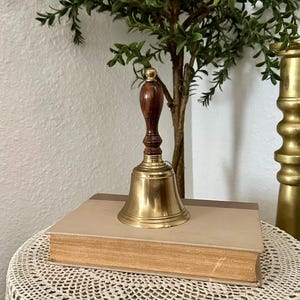 May include: A hand bell featuring a polished brass bell body and a dark brown wooden handle with a small brass finial at the top. The bell is resting on top of a closed, rectangular hardcover book with a light tan cover and visible page edges.