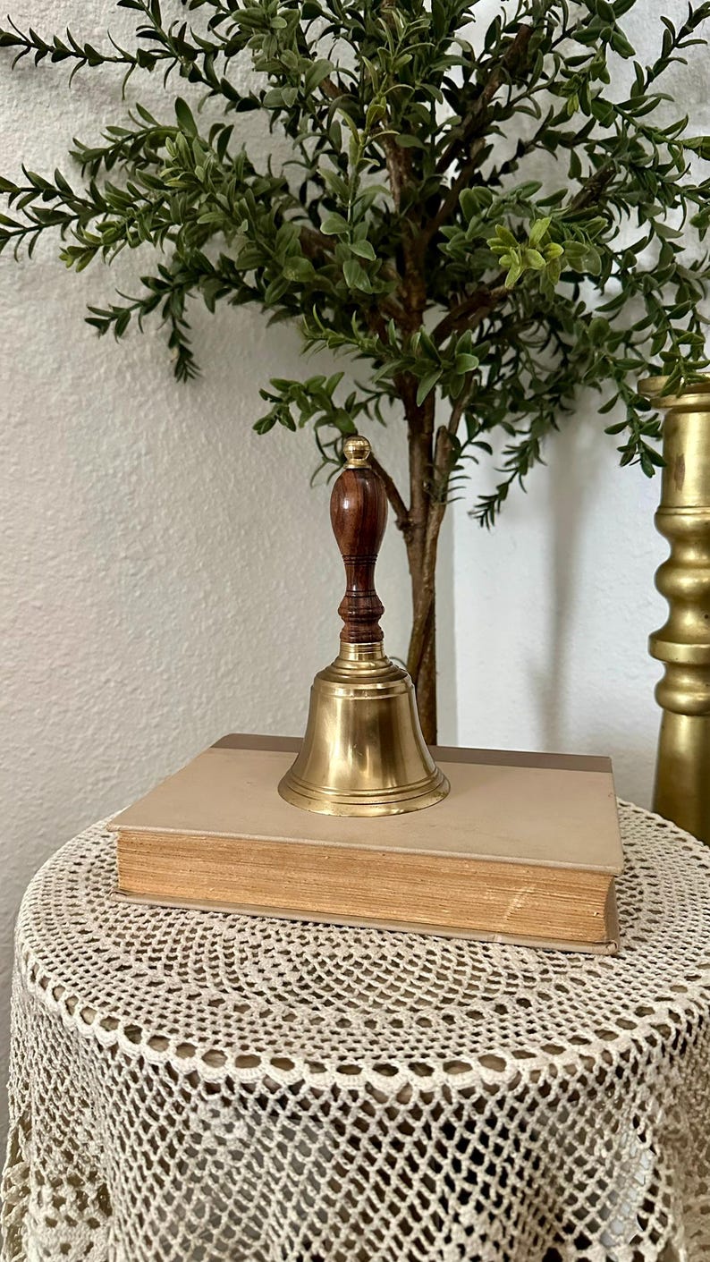 May include: A hand bell featuring a polished brass bell body and a dark brown wooden handle with a small brass finial at the top. The bell is resting on top of a closed, rectangular hardcover book with a light tan cover and visible page edges.