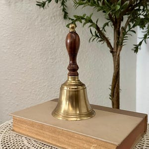 May include: A hand bell featuring a polished brass bell base and a dark brown wooden handle with a rounded top. The bell is positioned on top of a rectangular book with a light tan cover and visible pages along the side edge.