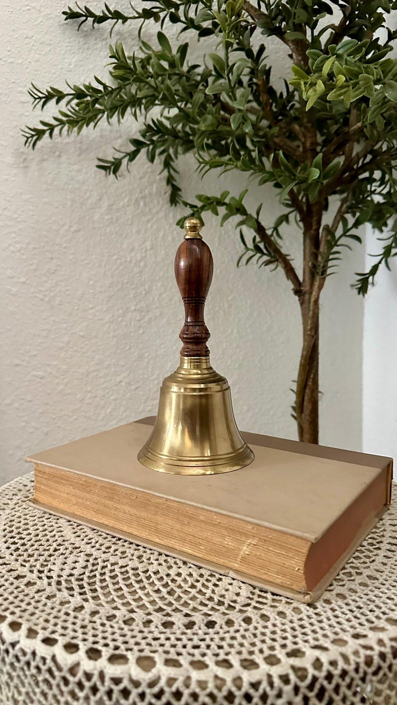 May include: A hand bell featuring a polished brass bell base and a dark brown wooden handle with a rounded top. The bell is positioned on top of a rectangular book with a light tan cover and visible pages along the side edge.