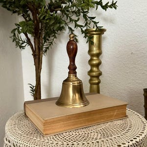 May include: A hand bell with a polished brass body and a dark brown wooden handle. The bell is positioned on top of a closed, rectangular book with a light tan cover. The handle features a small, rounded brass finial at the very top.