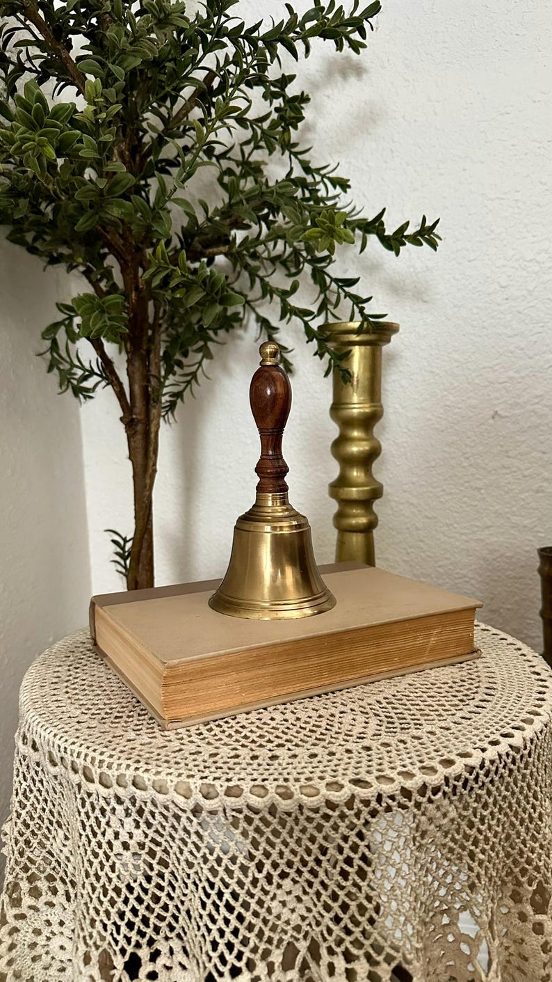 May include: A hand bell with a polished brass body and a dark brown wooden handle. The bell is positioned on top of a closed, rectangular book with a light tan cover. The handle features a small, rounded brass finial at the very top.