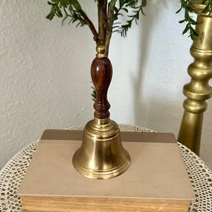 May include: A hand bell featuring a polished brass bell base and a dark brown wooden handle with a rounded top. The bell is positioned on top of a rectangular book with a light tan cover, showcasing a classic design with metallic and wood textures.