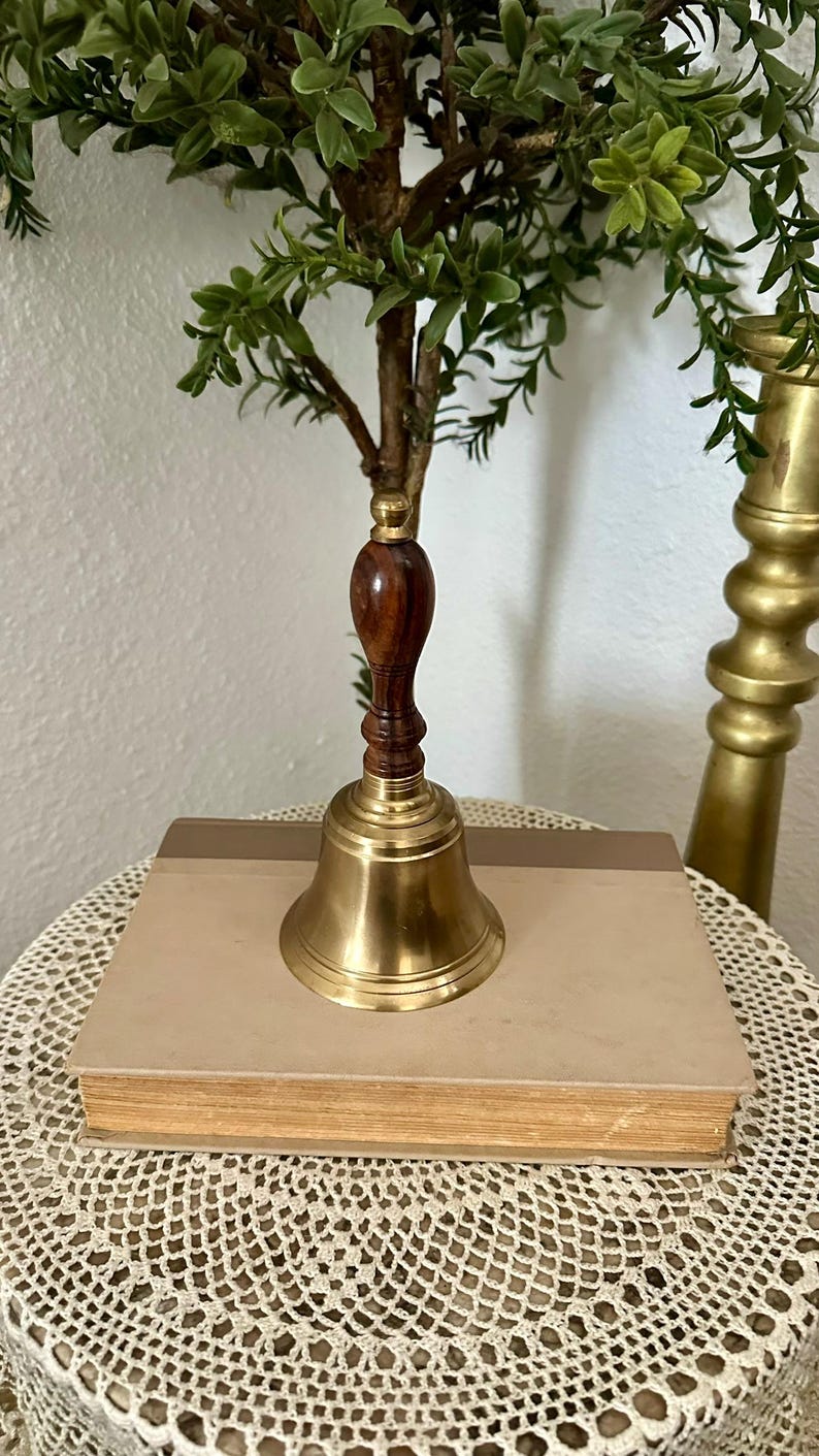 May include: A hand bell featuring a polished brass bell base and a dark brown wooden handle with a rounded top. The bell is positioned on top of a rectangular book with a light tan cover, showcasing a classic design with metallic and wood textures.