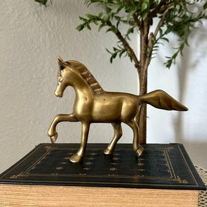May include: A polished brass horse figurine standing in a prancing pose with one front leg raised. The horse has a smooth, metallic gold-toned finish, a defined mane, and a flowing tail, capturing a classic equine form in a standing position.