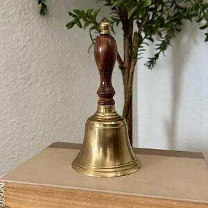 May include: A hand bell featuring a polished brass bell base and a dark brown wooden handle with a small brass finial at the top. The handle has a turned, ribbed design, and the brass bell has a smooth, flared rim and a reflective metallic finish.